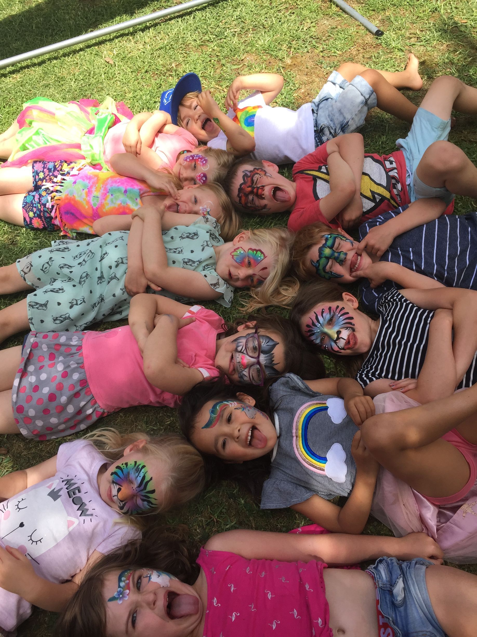 Children with face paint lie on the grass, smiling and laughing.
