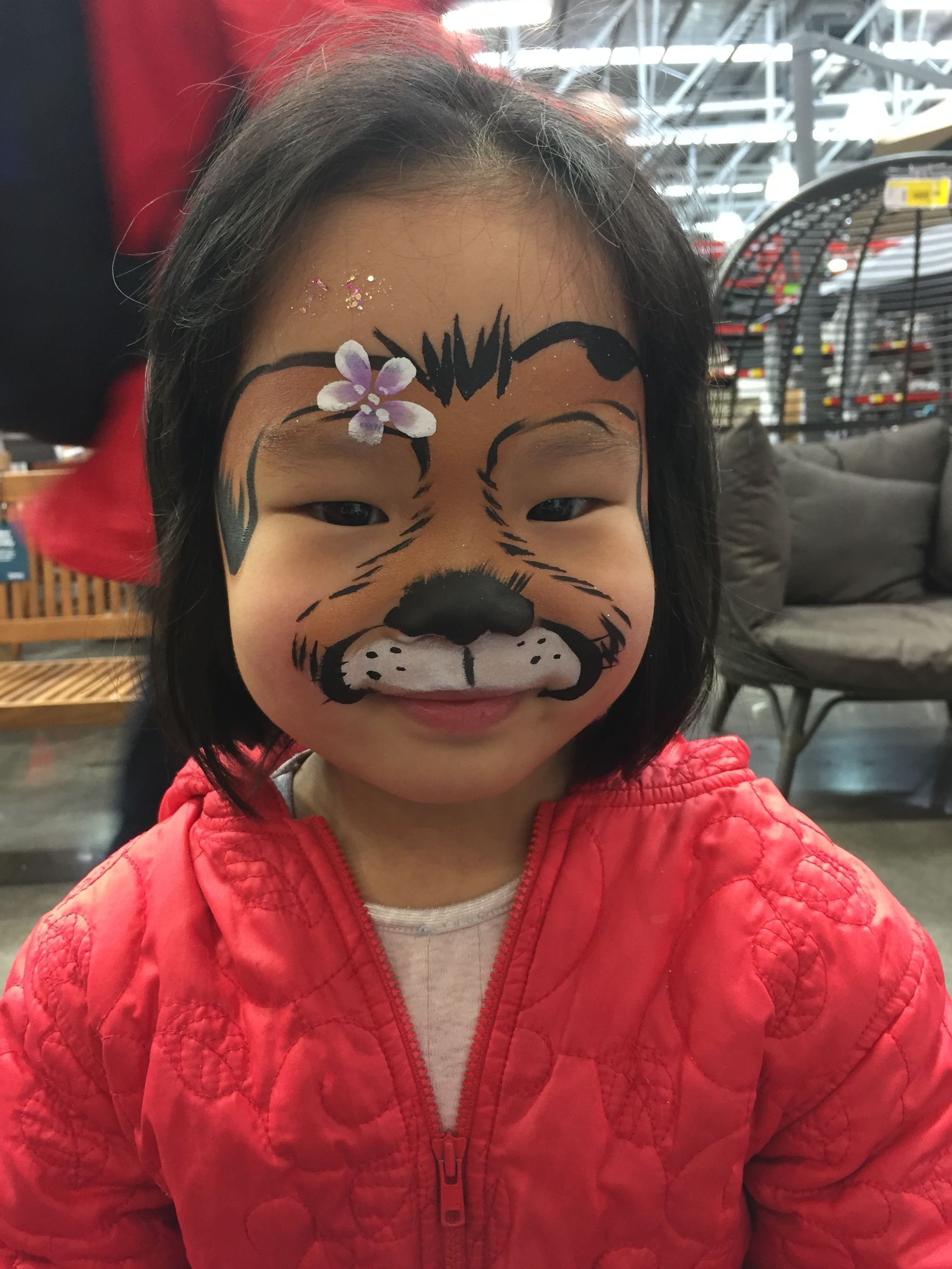 Child with face paint of a brown bear, wearing a red jacket, smiling.