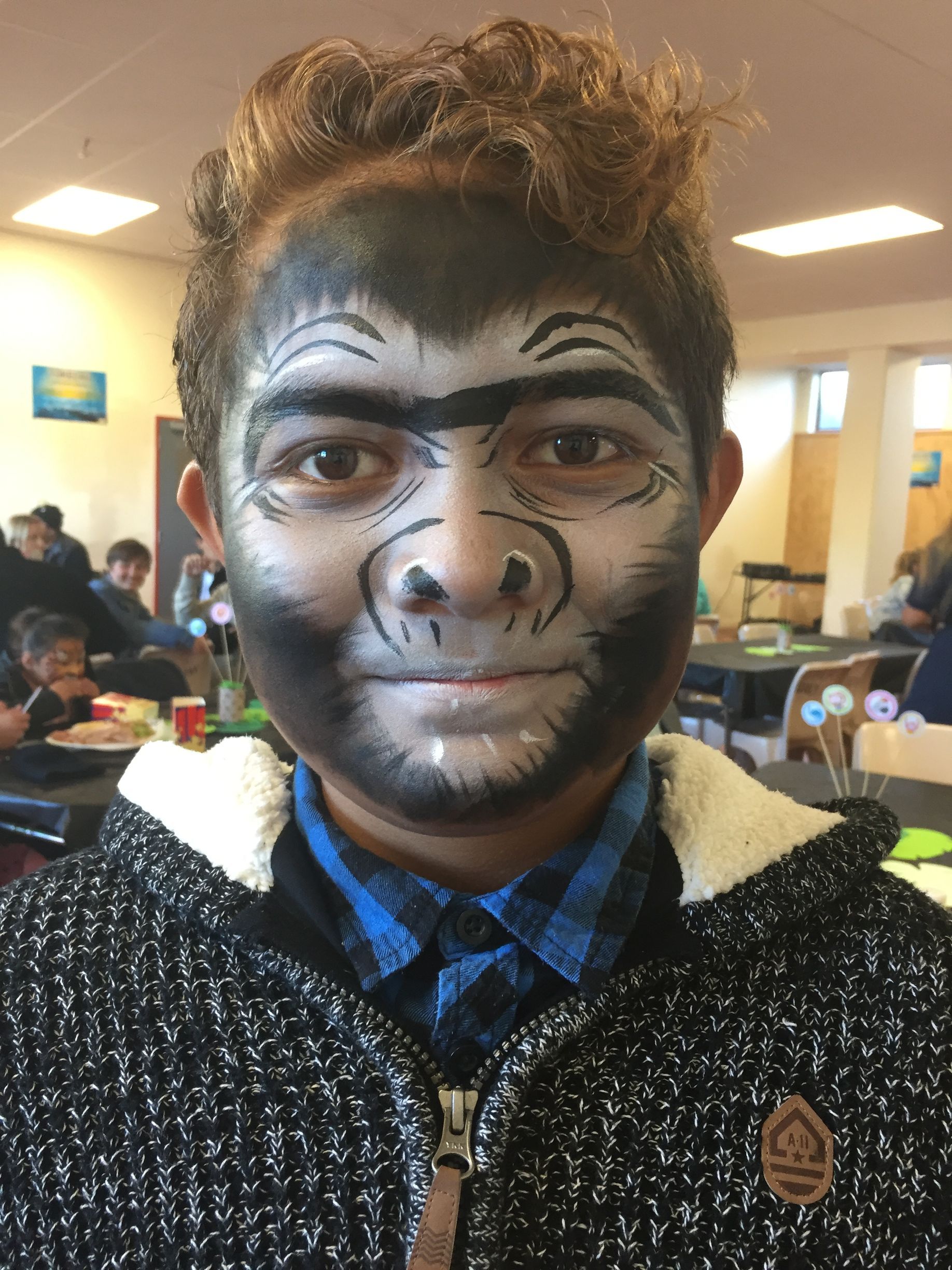 Boy with gorilla face paint, indoors. Black, white, and gray face paint, smiling, wearing a blue plaid shirt and gray jacket.