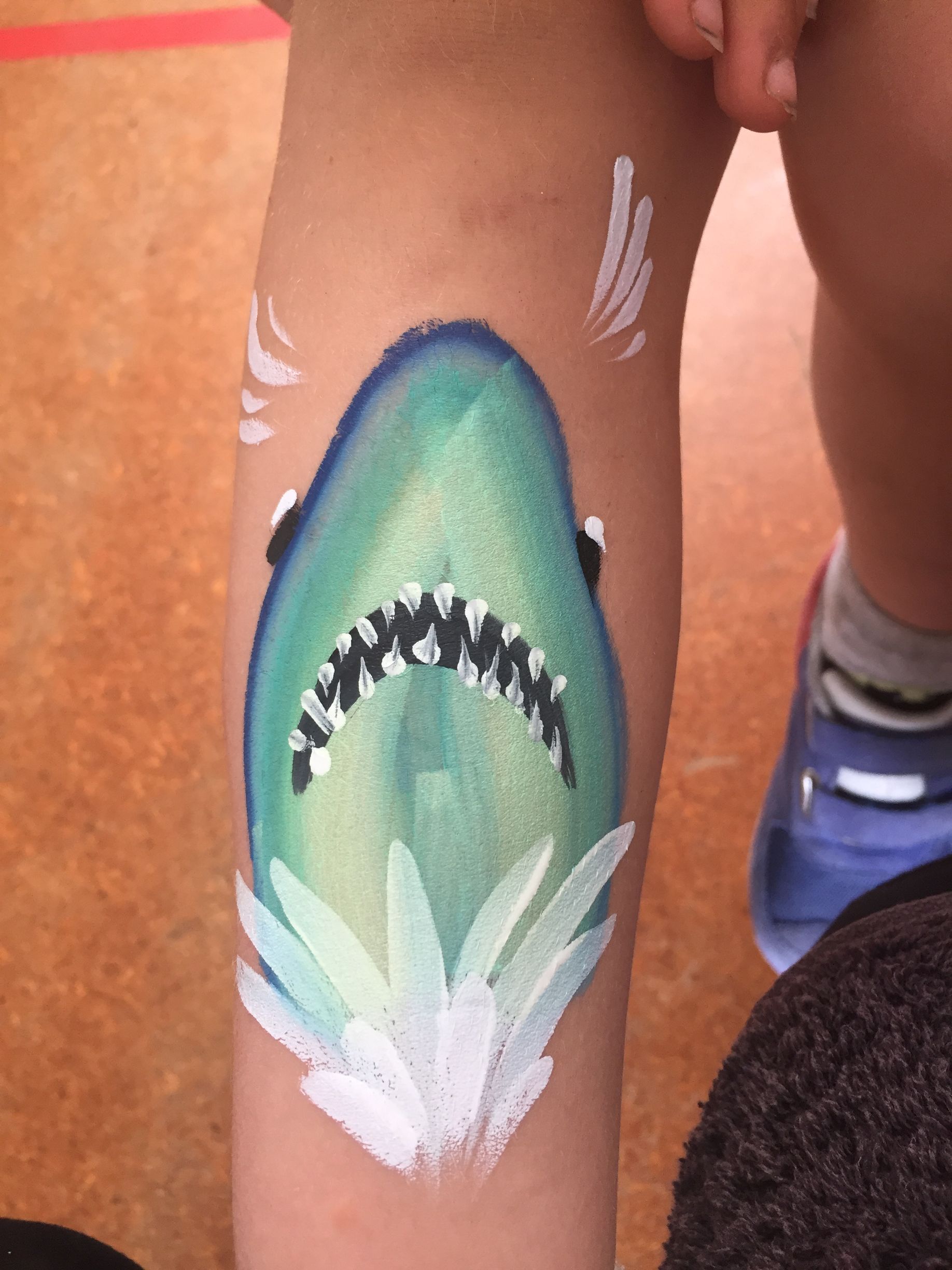 Shark face painted on a child's arm, blue, green, white, with open jaws and water effects.