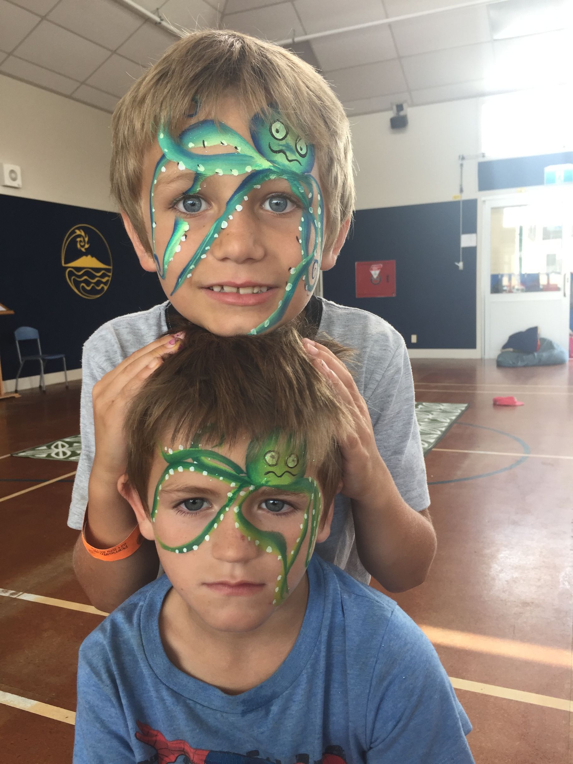 Two boys with green octopus face paint, one behind the other in a gymnasium.