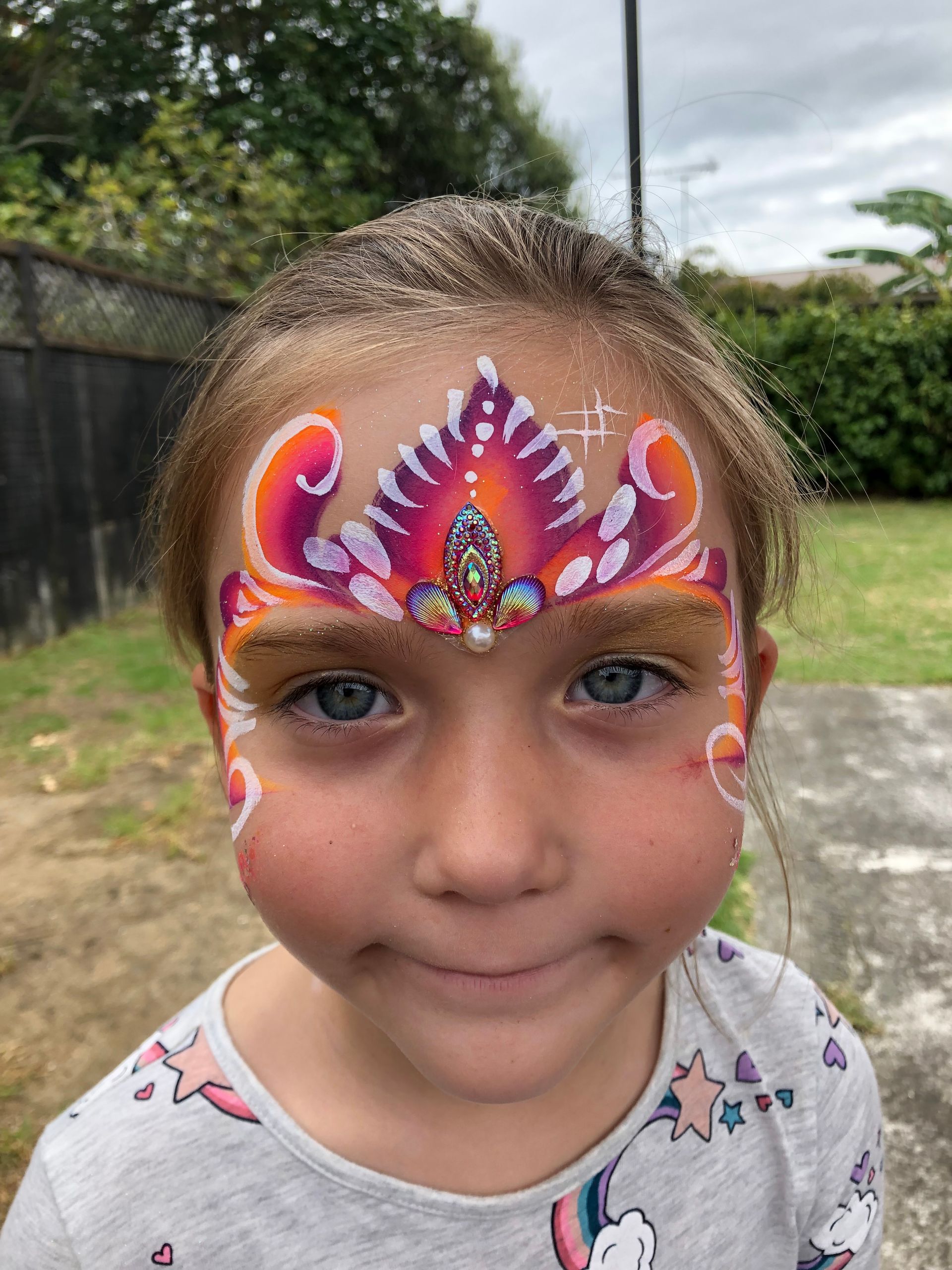 Girl with face painted as a pink and orange jeweled crown, set outdoors.