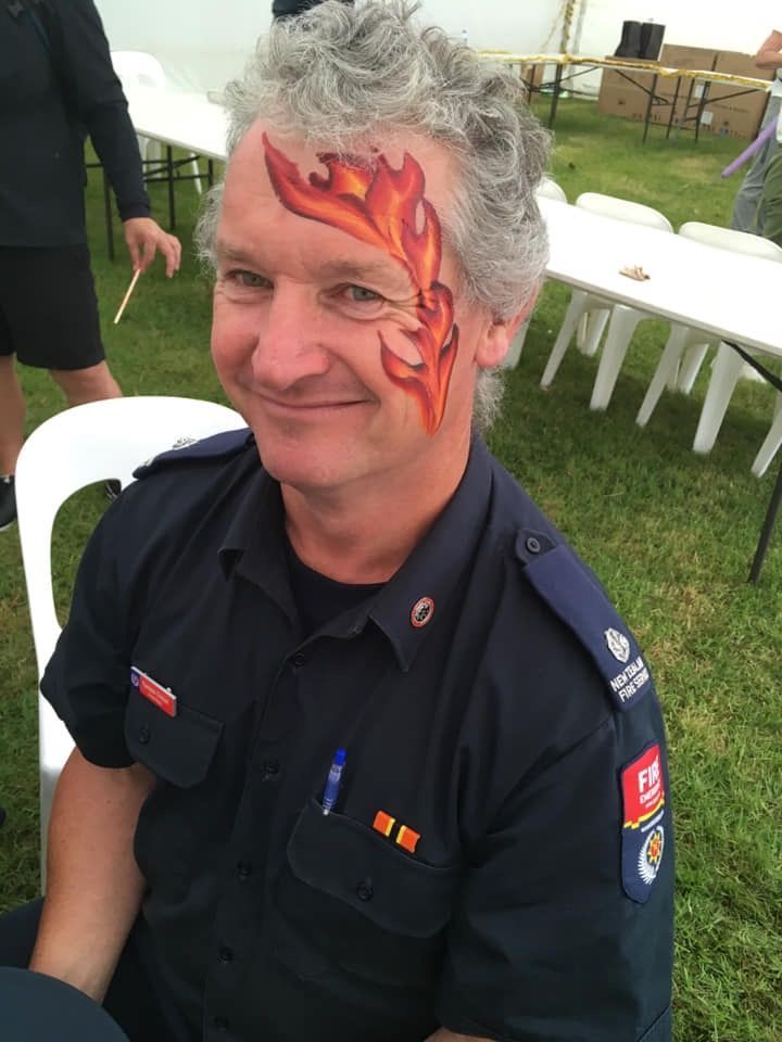 Man with grey hair and face paint (flame design), wearing a dark blue uniform, smiling outdoors.