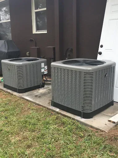 Two air conditioners are sitting on a sidewalk in front of a house.