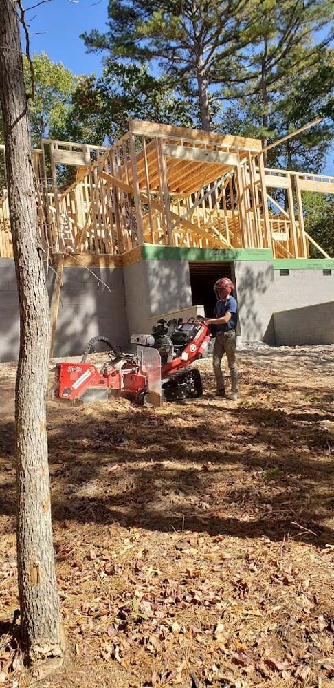 A man is standing next to a stump grinder in front of a house under construction.