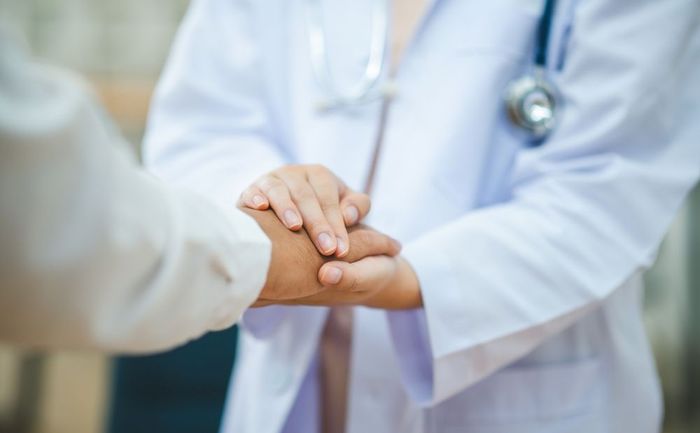 A doctor in a white coat with a stethoscope around their neck holds a patient's hand in a gesture of comfort.