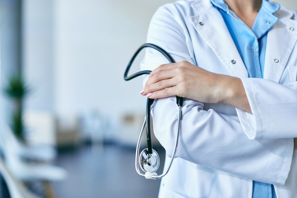 A professional in a white lab coat and blue scrubs standing with arms crossed, holding a stethoscope.