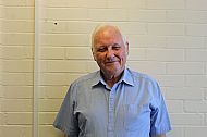 Man in a blue shirt smiles in front of a light-colored brick wall.