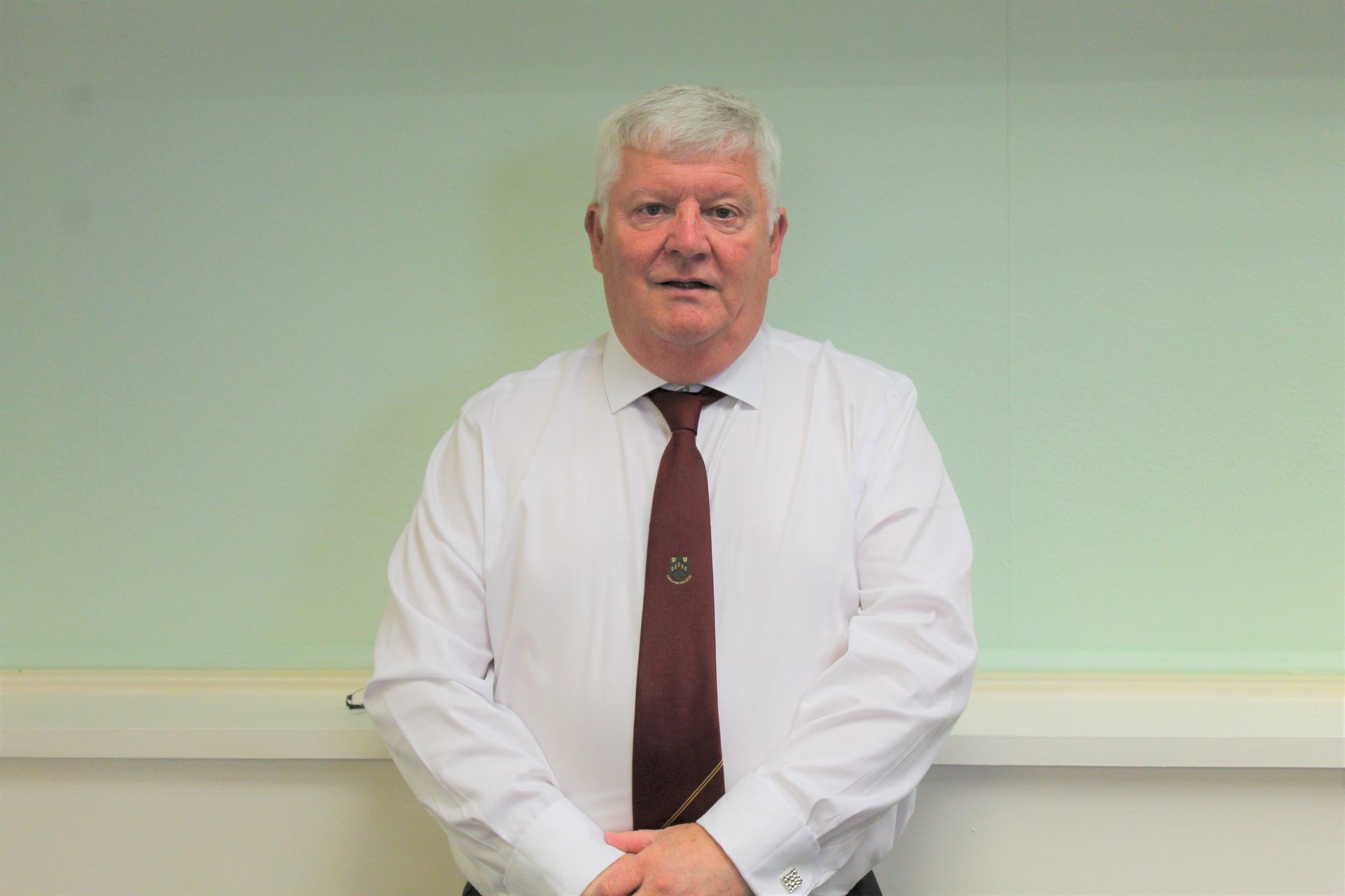 Man in white shirt and maroon tie, standing in front of a light green wall.
