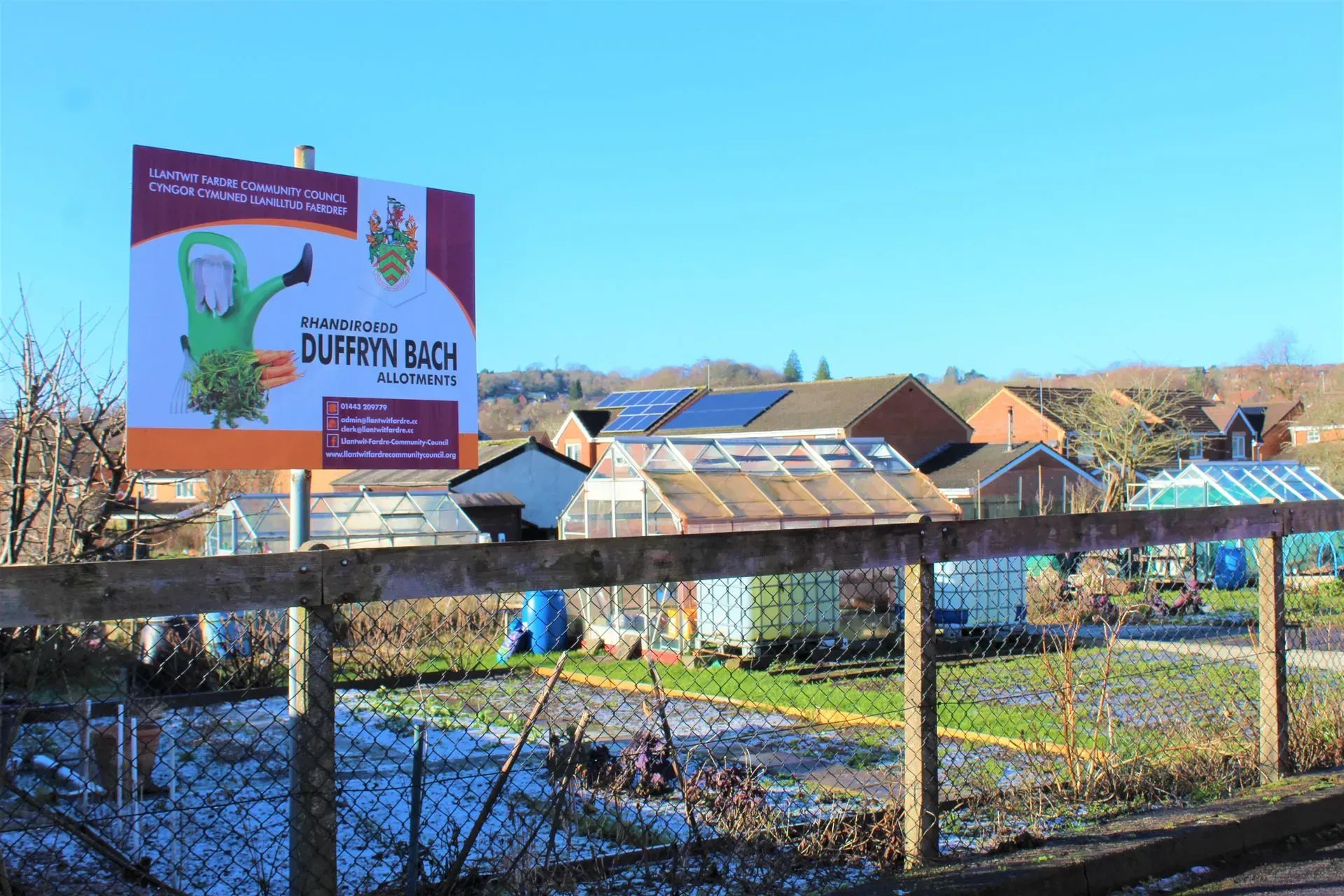 Billboard in front of allotment gardens with a cartoon figure holding a thumbs up. Buildings and blue sky in background.