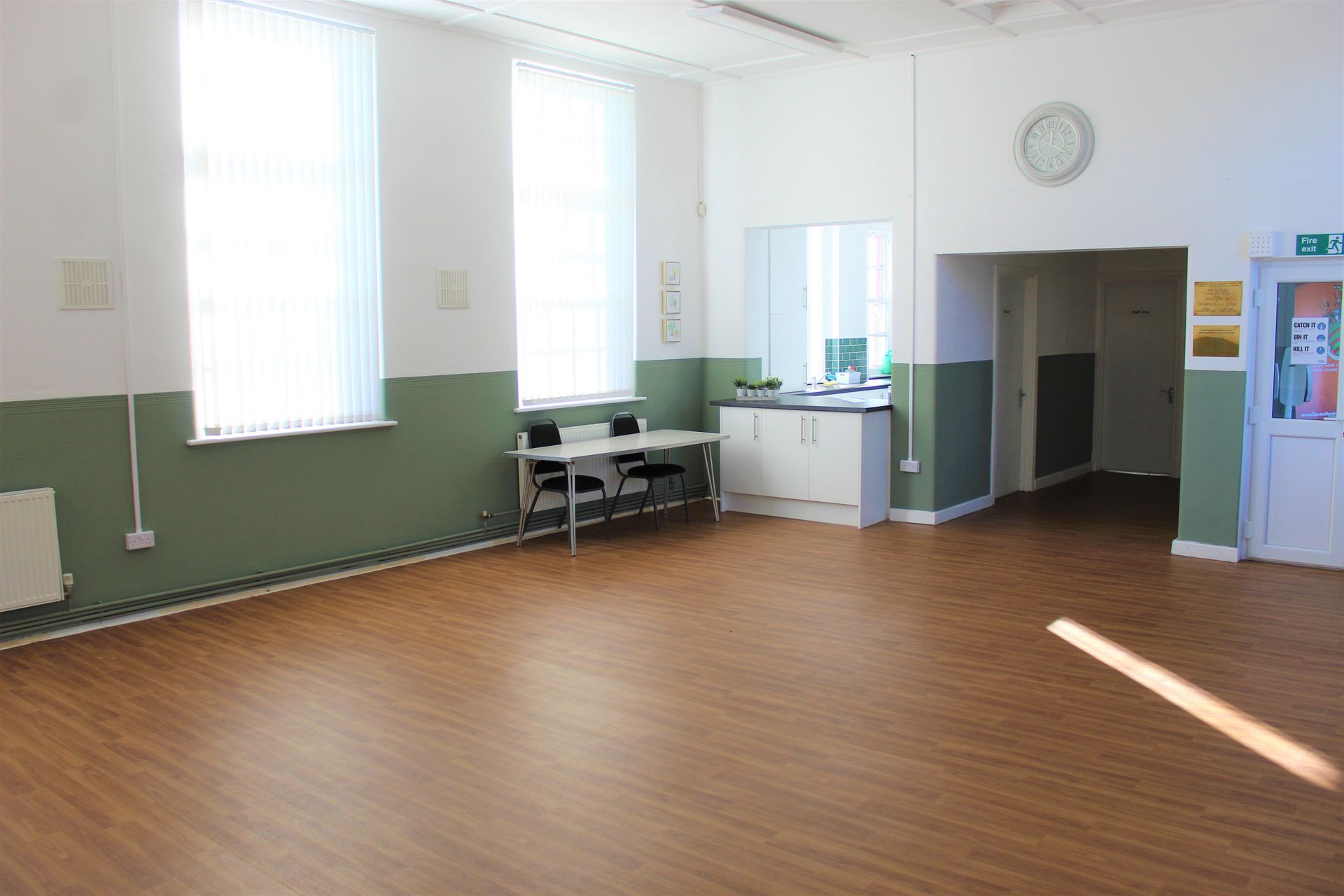 Empty hall with wooden floor, two windows, and green-painted lower walls. White cupboards and door.
