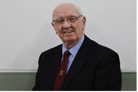 Smiling man in suit, glasses, and tie, standing against a white wall.
