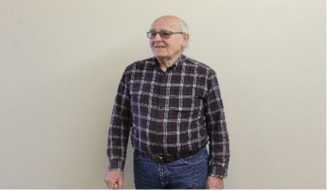 Older man wearing glasses and a plaid shirt stands in front of a neutral wall.