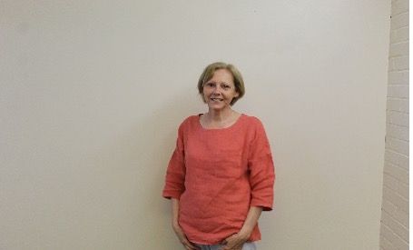 Woman in an orange shirt smiles, standing against a beige wall.
