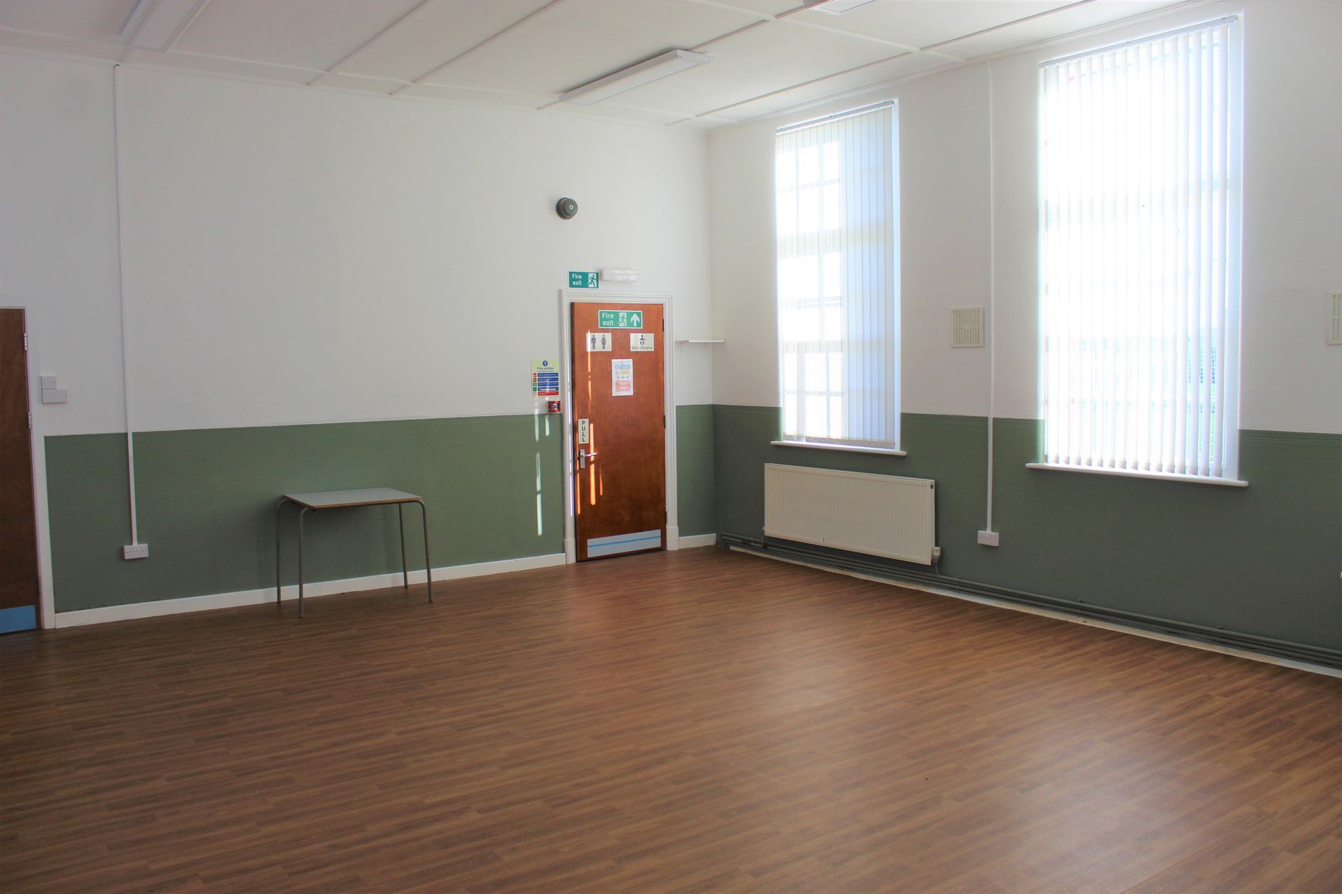 Empty community hall with wood floor, green and white walls, door, windows, and small table.