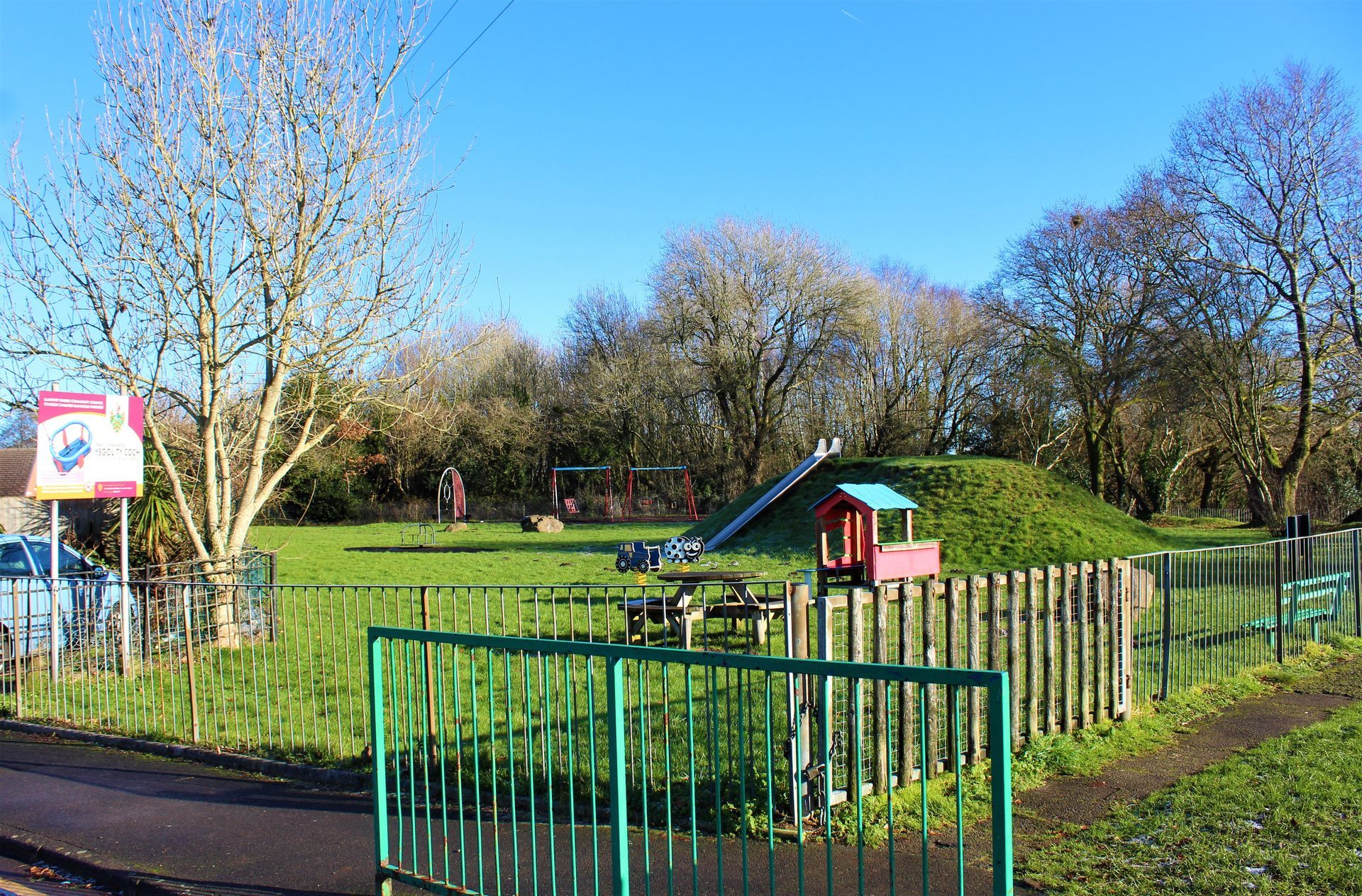 Green park with playground equipment behind a green fence under a blue sky.