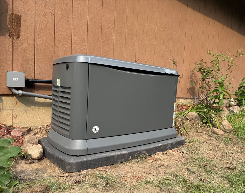 Gray home generator next to a brown house, on a dark foam base, outdoors.