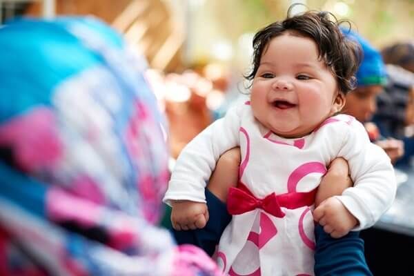 Smiling baby in a white shirt with a pink bow, being held at an outdoor gathering