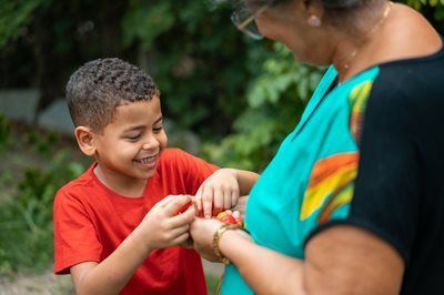 Child in red shirt smiling while tying a bracelet on an adult's wrist outdoors