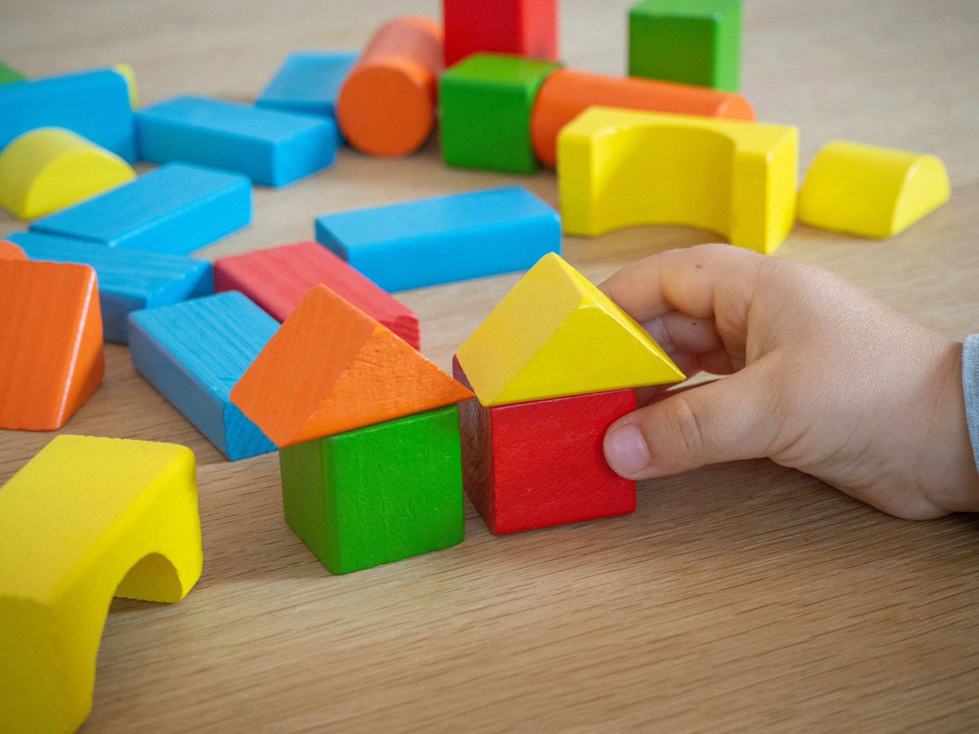 A hand places a yellow triangular block on a red square block, next to a small house made of a green cube and orange roof.