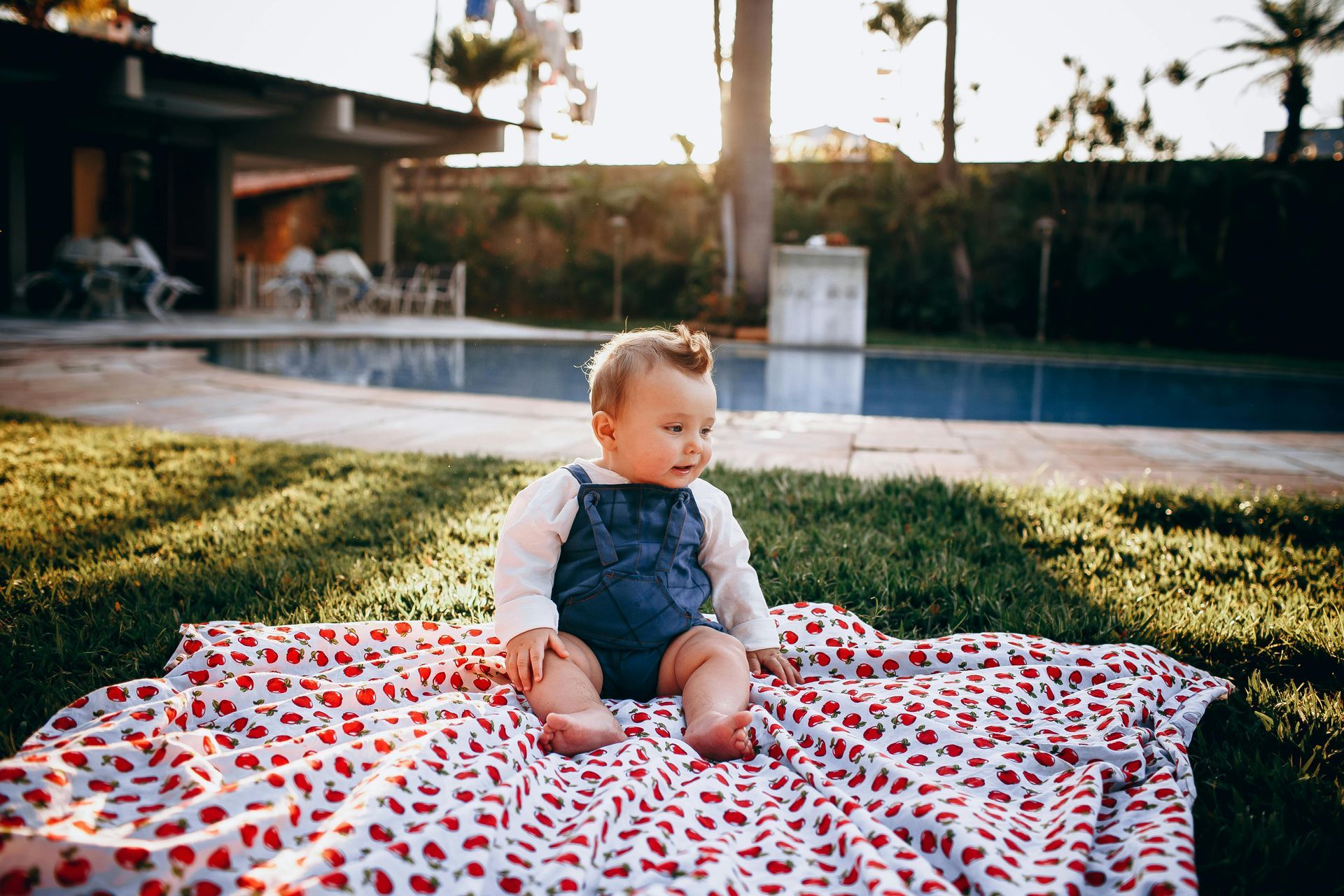 Baby sitting on a patterned blanket by a swimming pool at sunset.