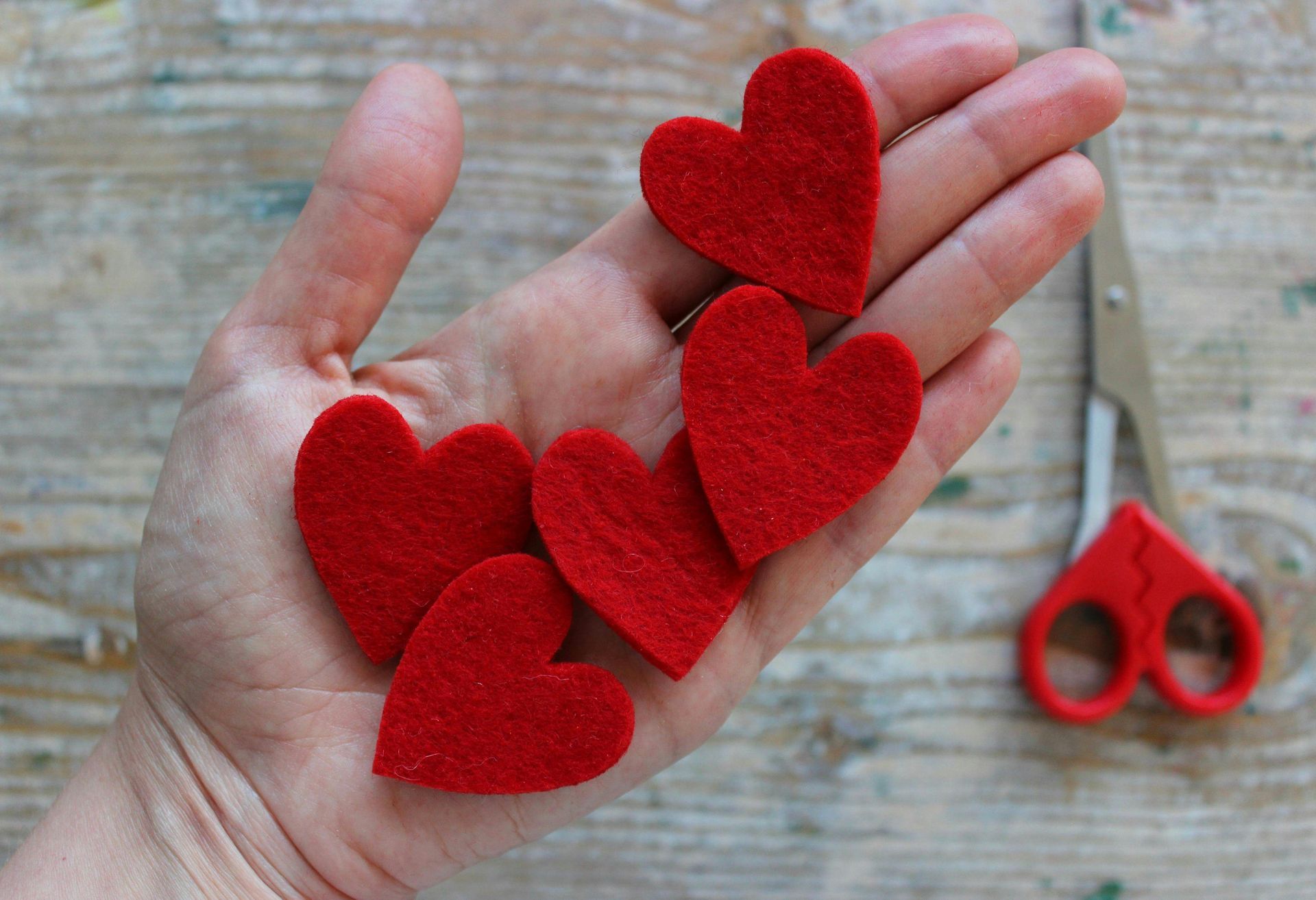 Hand holding five red felt hearts, scissors on wood surface.