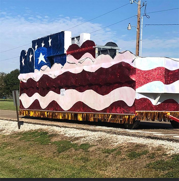 A parade float decorated as the American flag, red, white, and blue, parked on a gravel road.