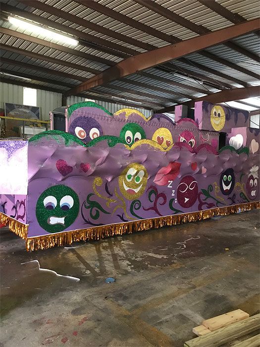 Purple float decorated with various emoji faces and gold fringe, in a large warehouse.