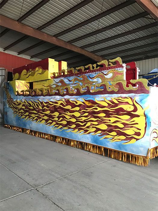 Mardi Gras float with flame-like designs in yellow, red, and blue. Gold fringe along the bottom in a warehouse.