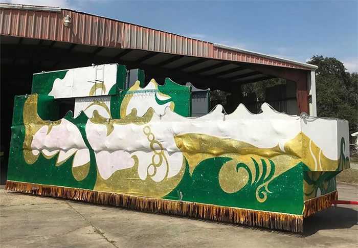 Mardi Gras float with green, gold, and white decorations, parked outside a building.