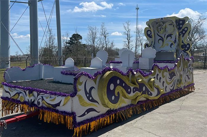 Mardi Gras float with white, gold, and purple decorations on a trailer, parked on asphalt.
