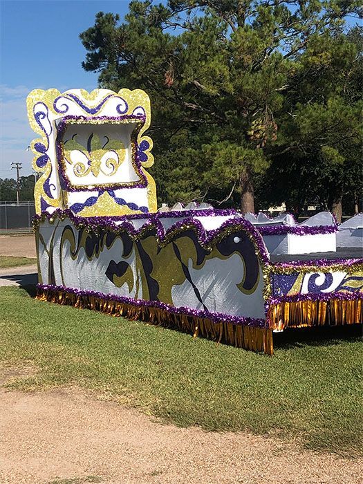 Mardi Gras parade float, purple, gold, and white, in a grassy area with trees.