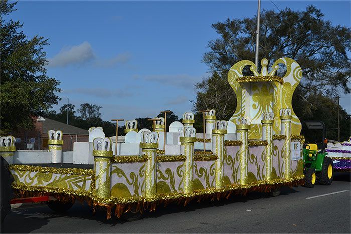 Mardi Gras parade float with gold and white decorations, pulled by a green tractor, on a sunny day.
