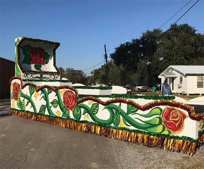 Mardi Gras float with large rose details, green and gold embellishments, parked on a street.