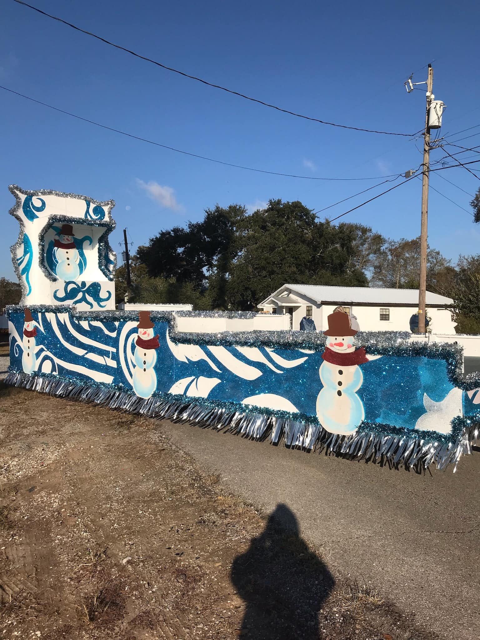 Snowman-decorated parade float with blue and white decorations, silver tinsel, and a snowman on top under a clear, sunny sky.