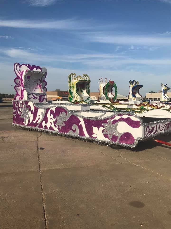 Mardi Gras float, white with purple and silver decorations, parked on pavement under a blue sky.