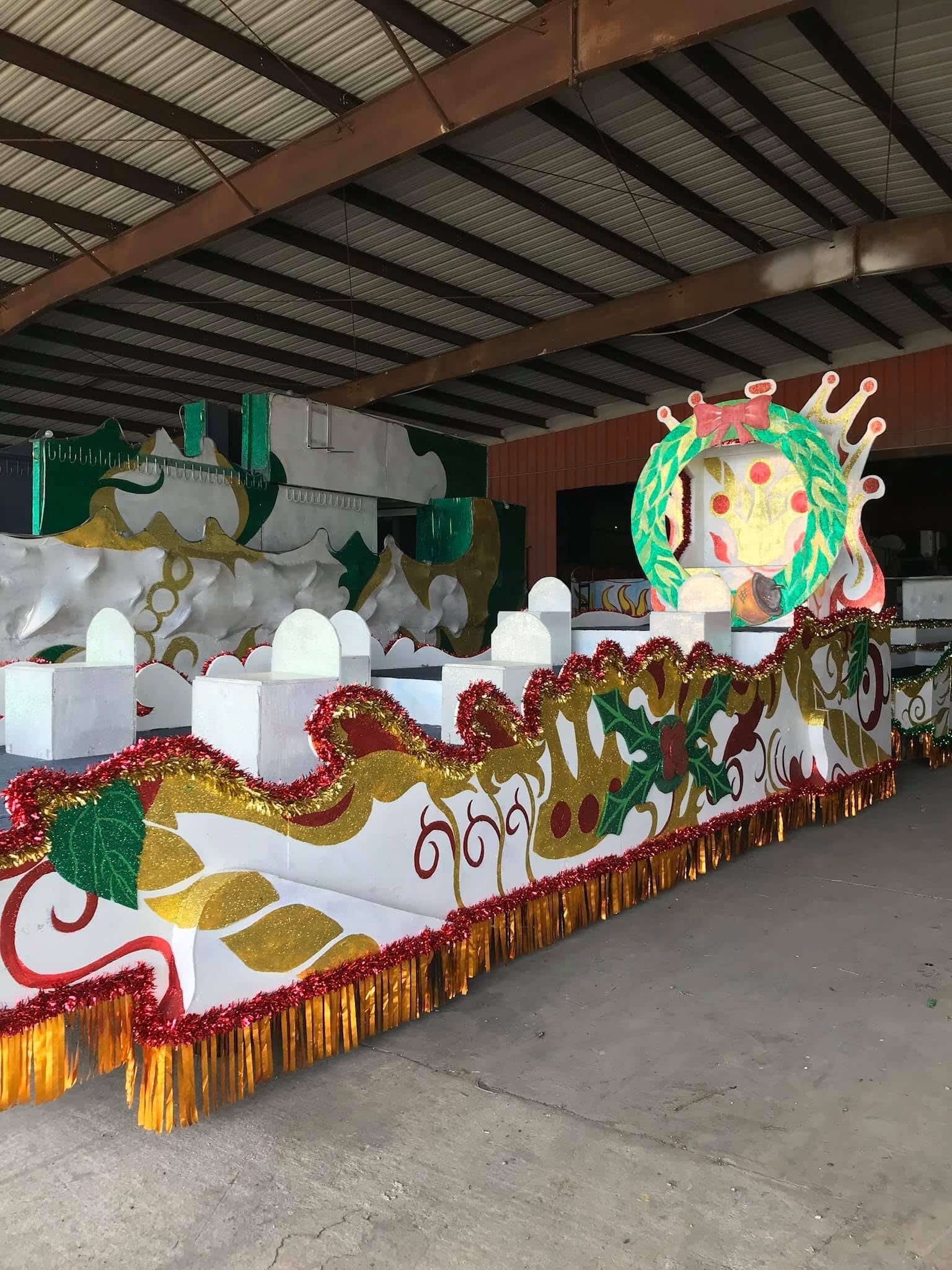 Mardi Gras float with white and gold decorations, inside a warehouse. Green wreath and figure on top.