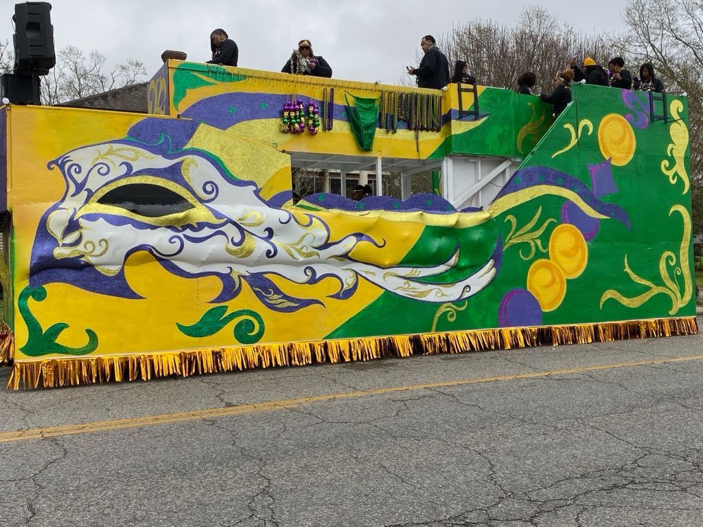 Mardi Gras parade float, decorated in glittery green, purple, and gold, parked in a grassy area with trees.