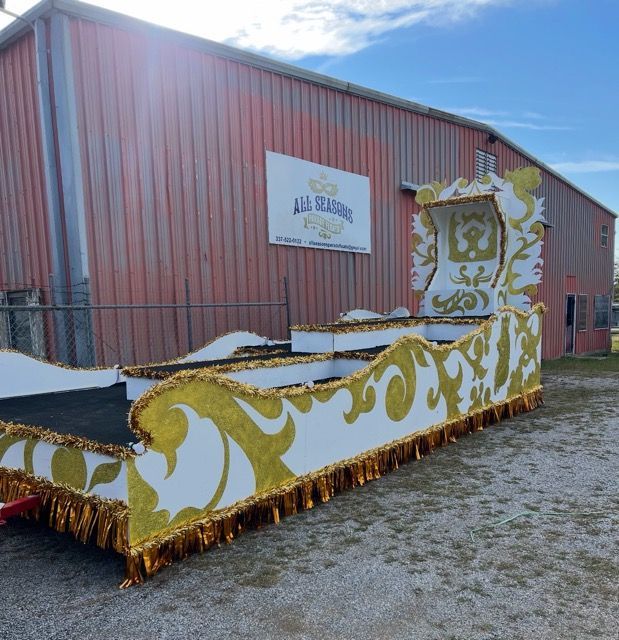 A parade float, white and gold with ornate designs, sits outside a red industrial building.