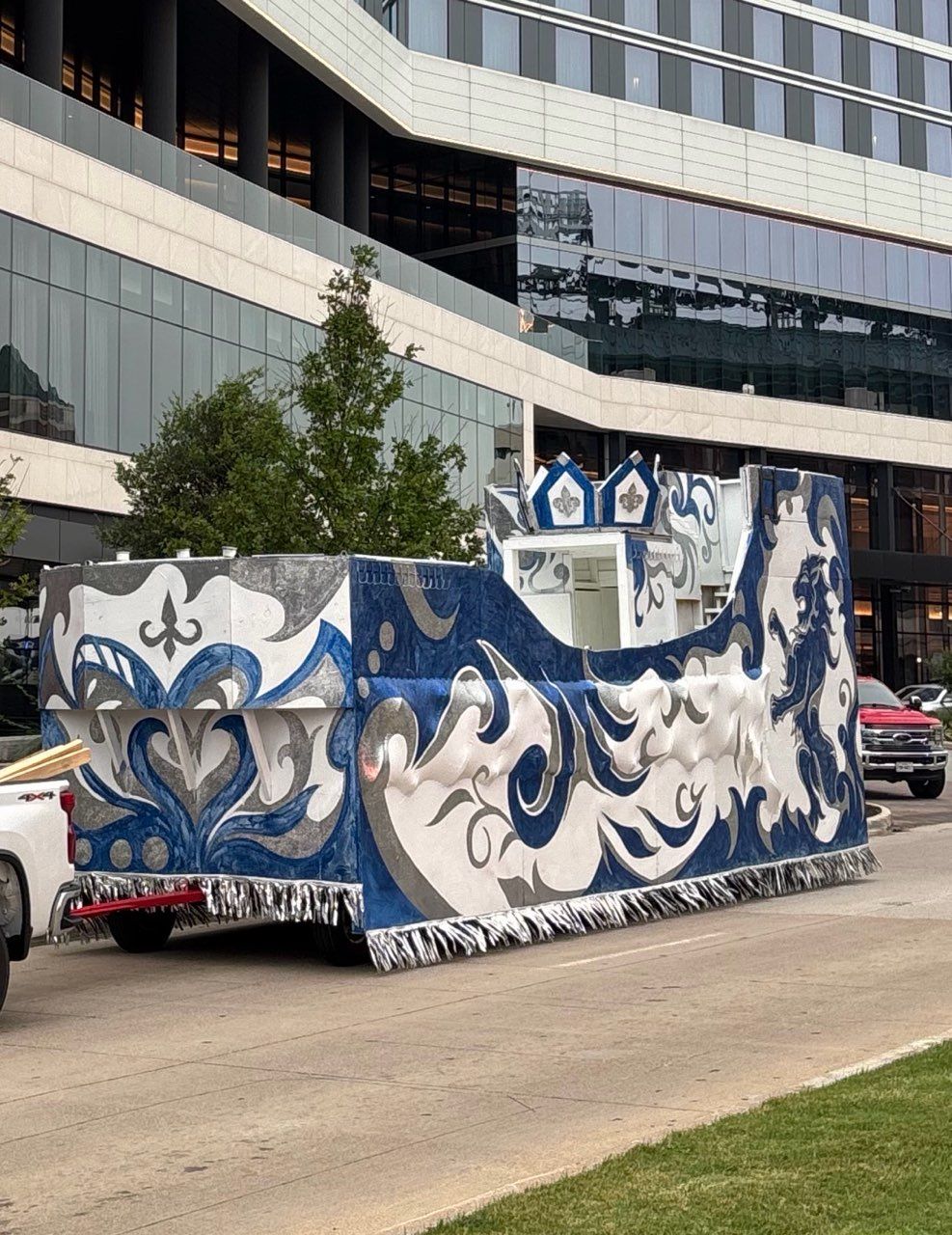 A parade float decorated in blue and white scrolls, pulled by a white truck, on a city street.