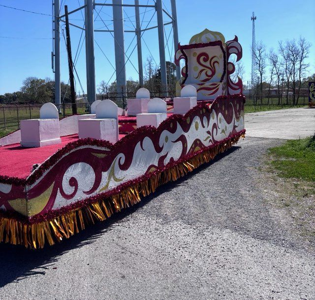 A brightly decorated parade float, with gold and red accents, parked on a gravel road, water tower in background.