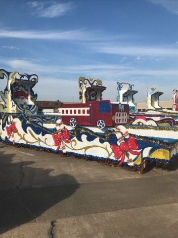 Colorful parade float with a red train, decorated with bows and blue waves, against a blue sky.