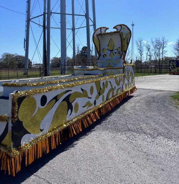 Mardi Gras float with gold and white decorations, parked on a road near a water tower.