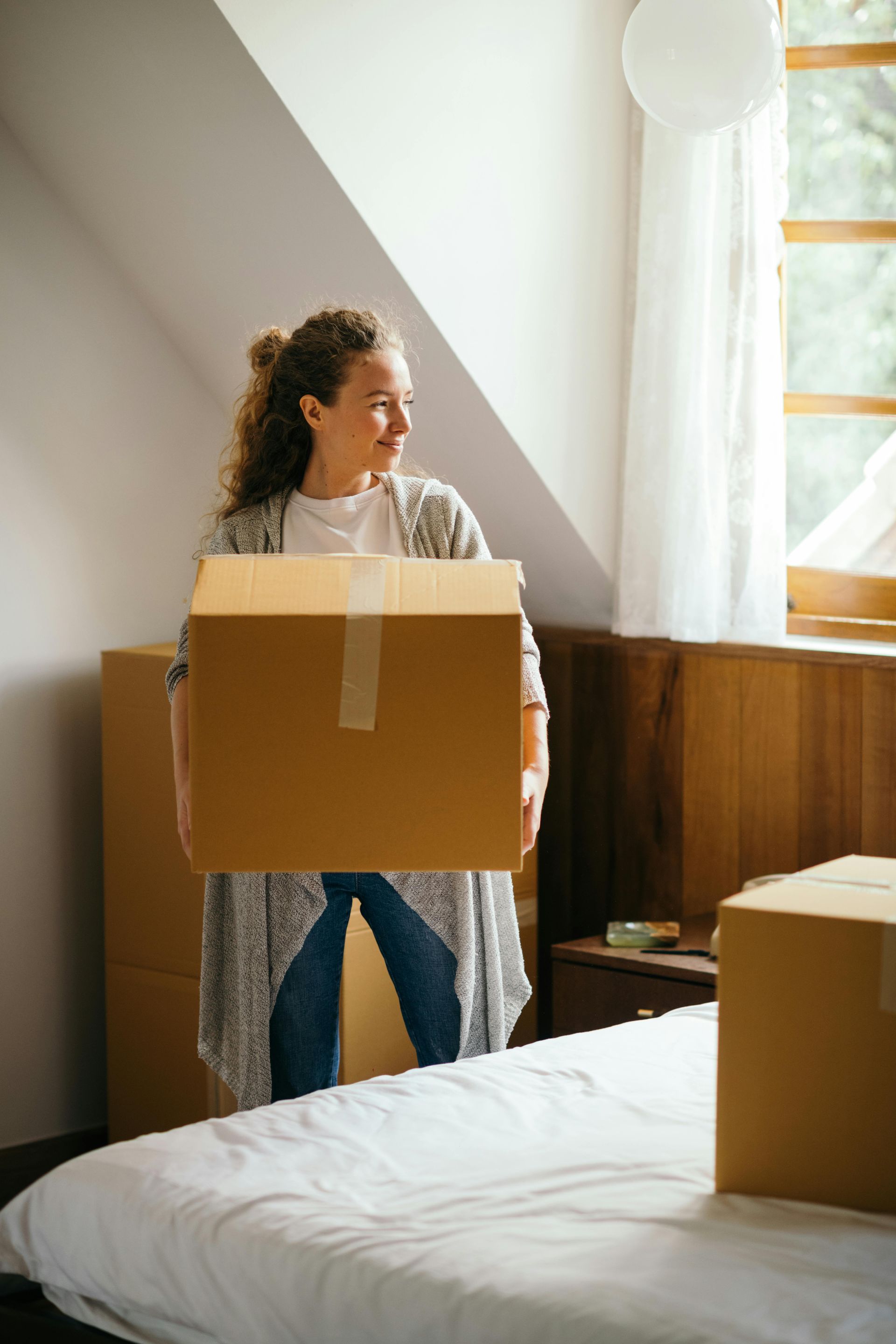 A woman is carrying a cardboard box in a bedroom.