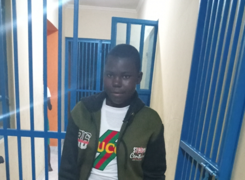 A young Black person stands in front of blue bars. They wear a white shirt with a green and black jacket.