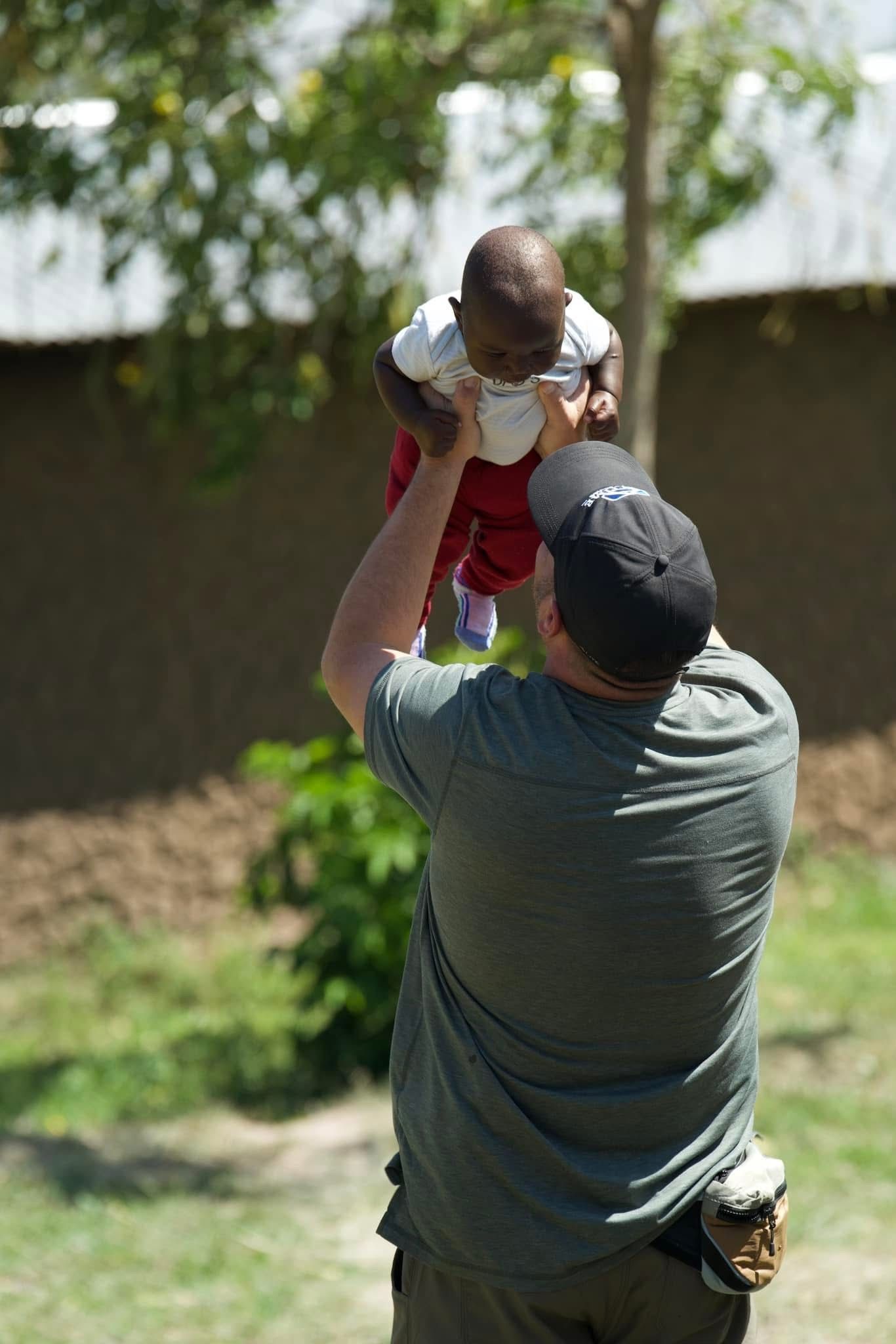 Man holding a baby up in the air outside. The baby wears a white shirt and red pants.