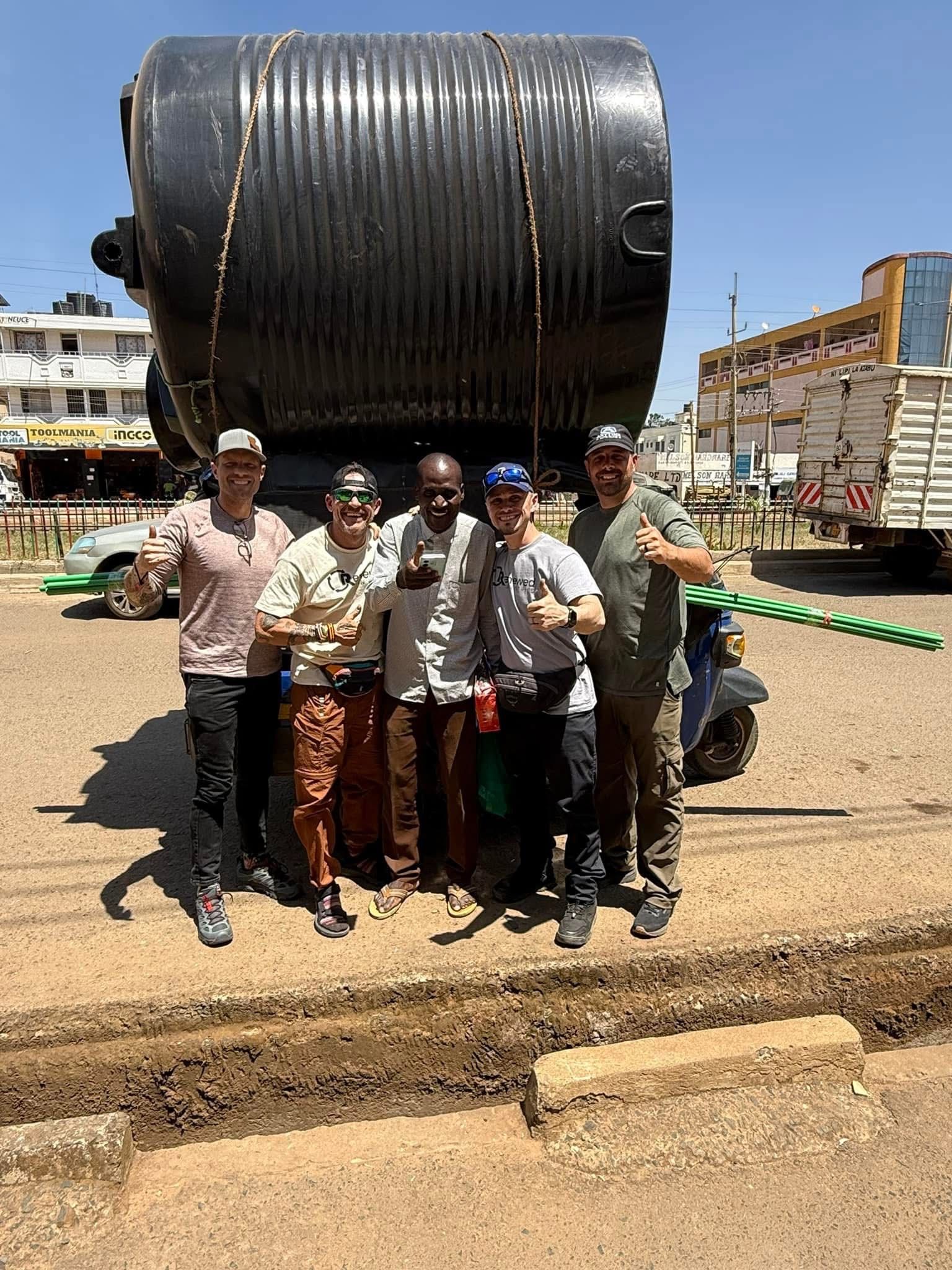 Group of six people posing in front of a large black tank on a small vehicle in a street, sunny day.