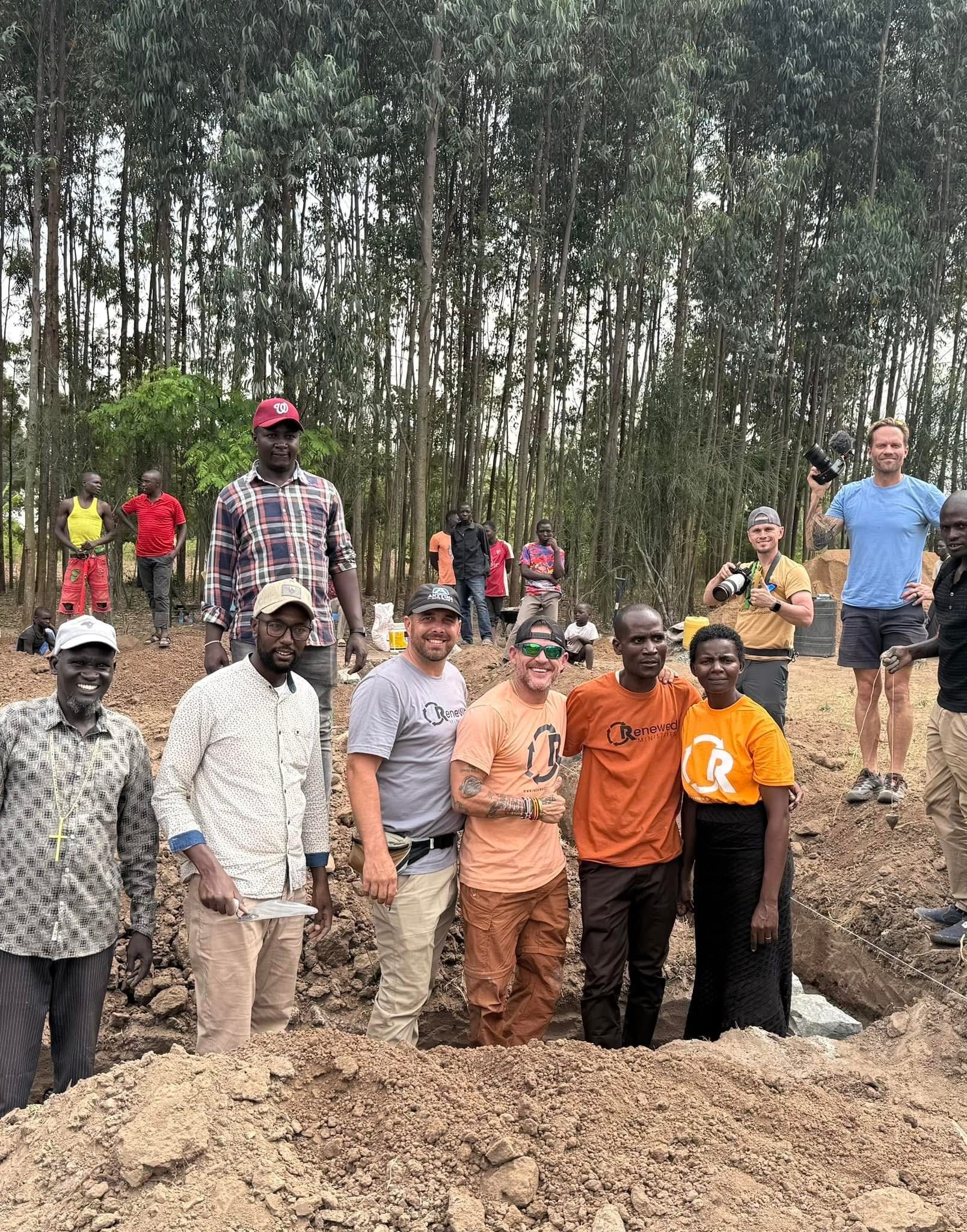 Group of people posing by a dug trench outdoors, near a line of tall trees; some in orange shirts.