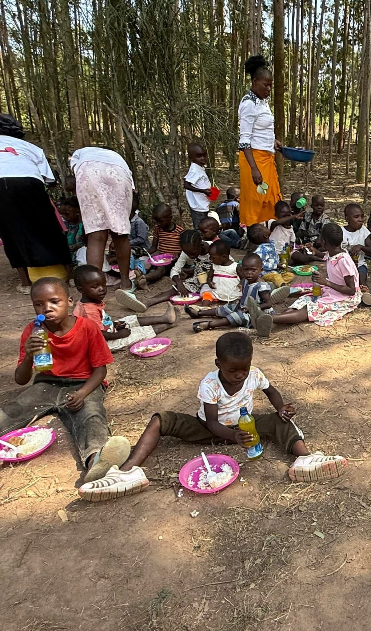 Children eating outdoors, some seated, some standing. Adults supervise. Forest background.