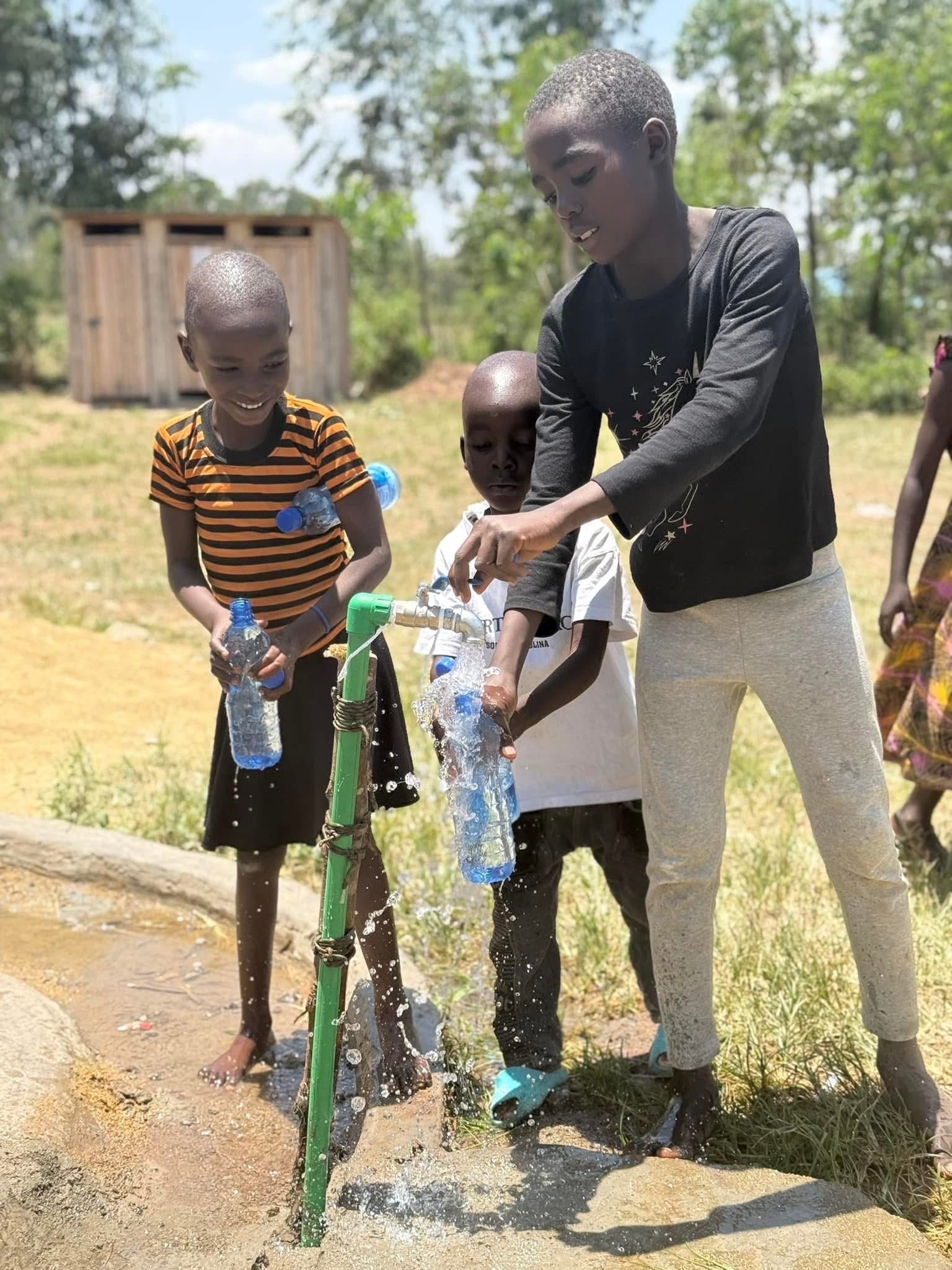 Children filling water bottles at an outdoor tap. They smile and play with the water. Background of trees and a building.